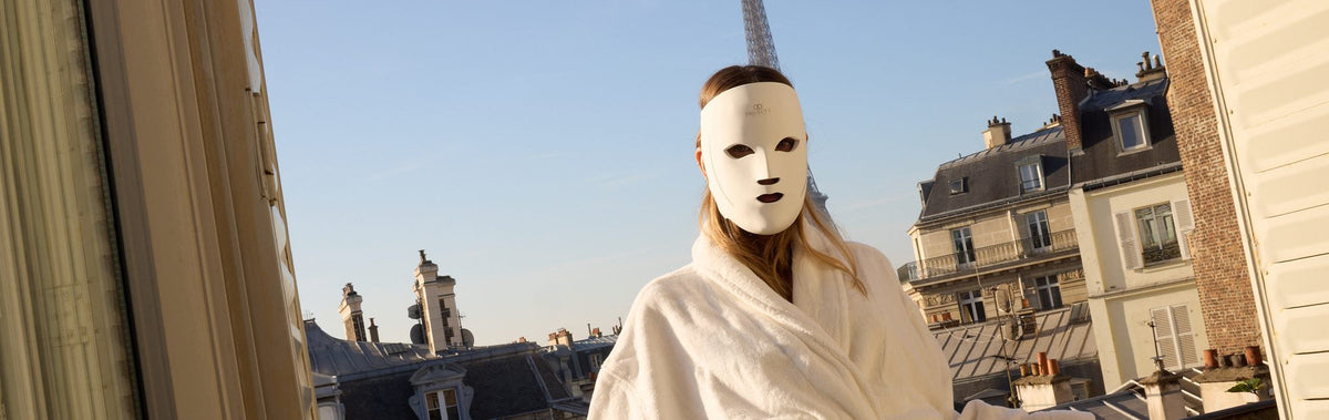 Woman wearing the LumaLux Face V2 LED mask leaning on a Paris balcony railing at sunrise